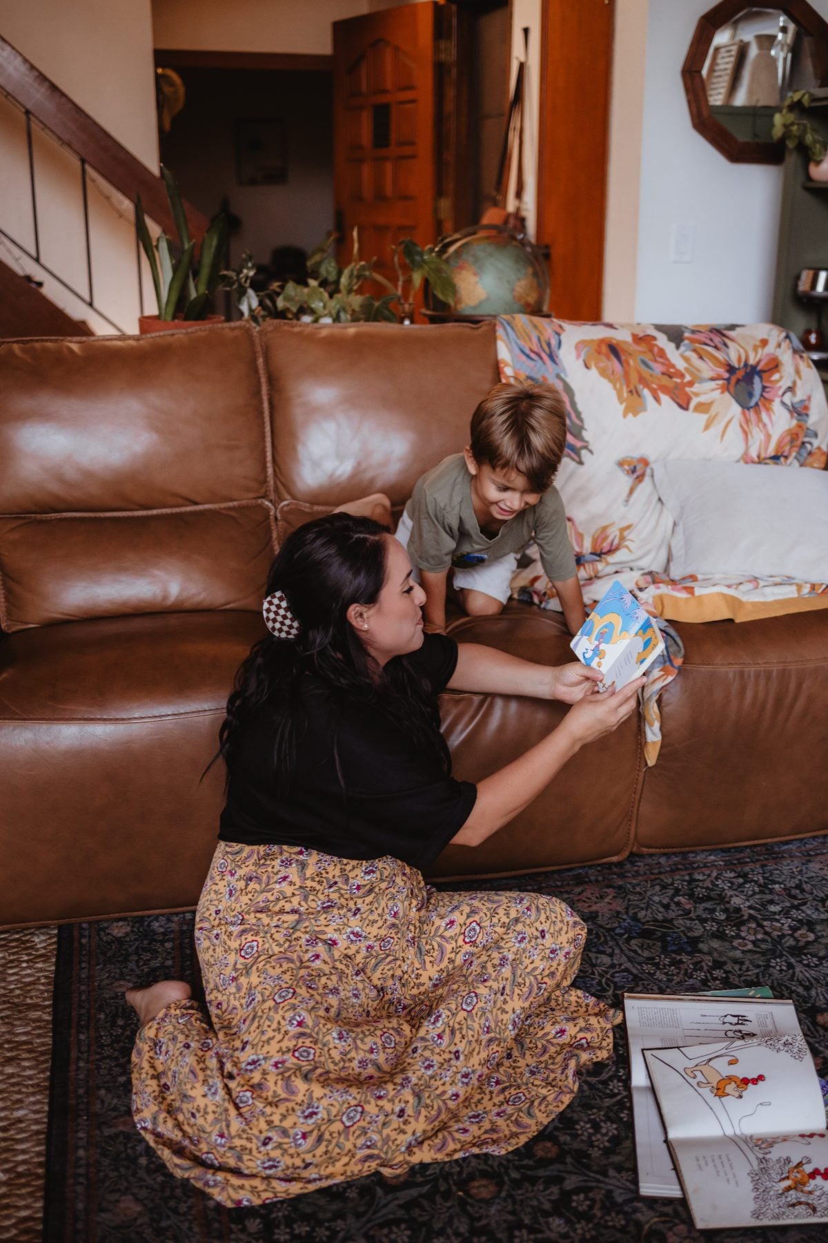 Mother hands storybook to toddler son beside leather sofa