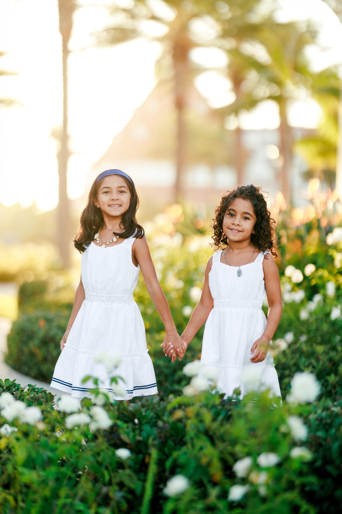 Sisters in white dresses hold hands among blooming white roses