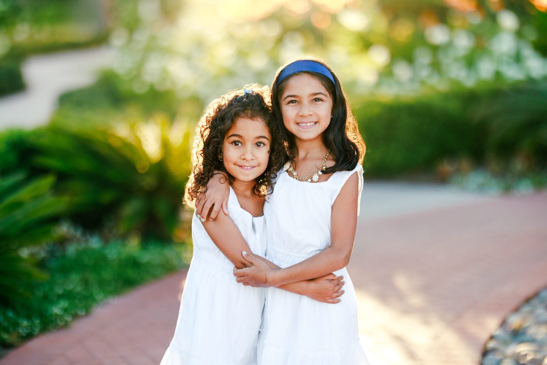 Two sisters in white dresses hug each other on a garden path