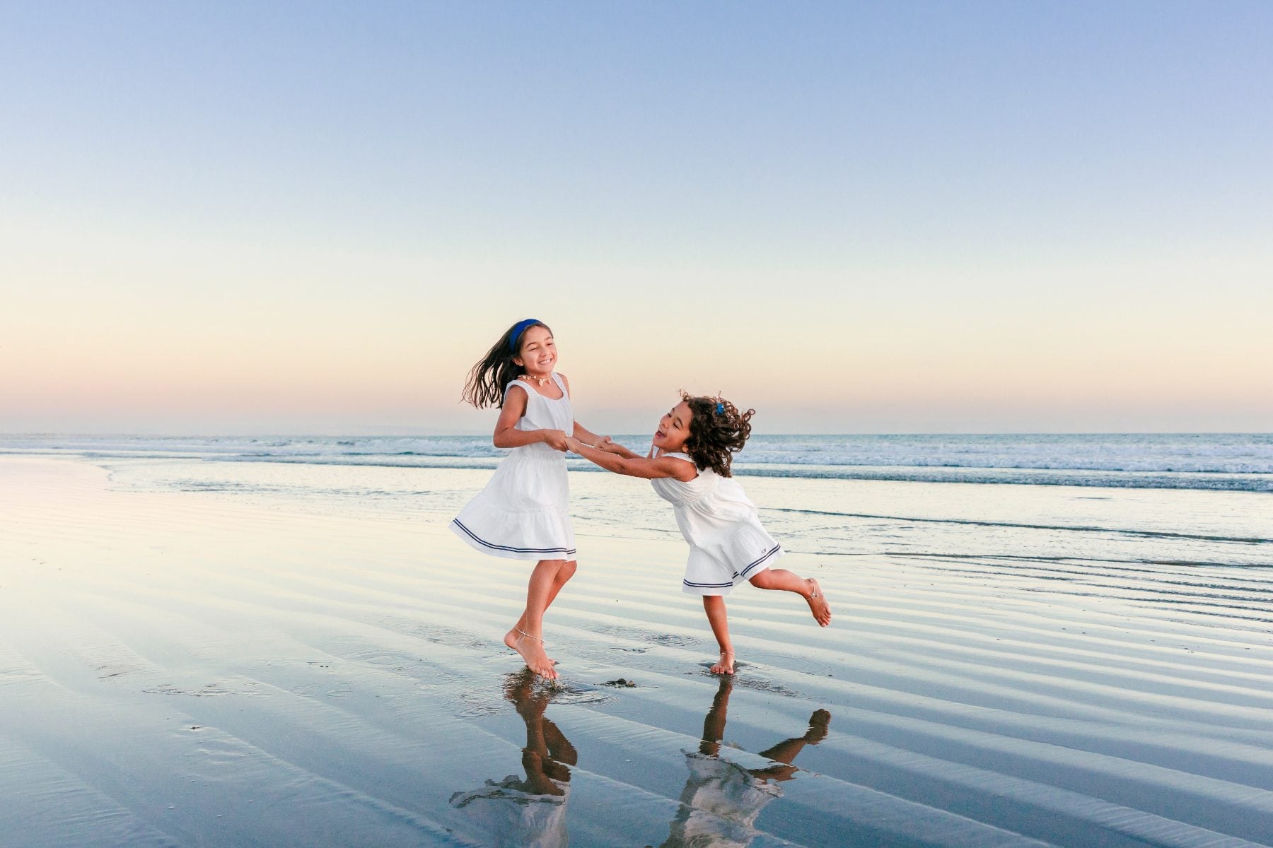 Two sisters in white dresses twirling and laughing on Coronado Beach