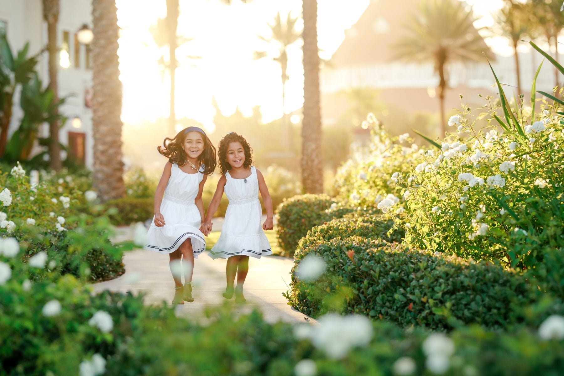 Two sisters in white dresses run through garden path at Hotel del Coronado