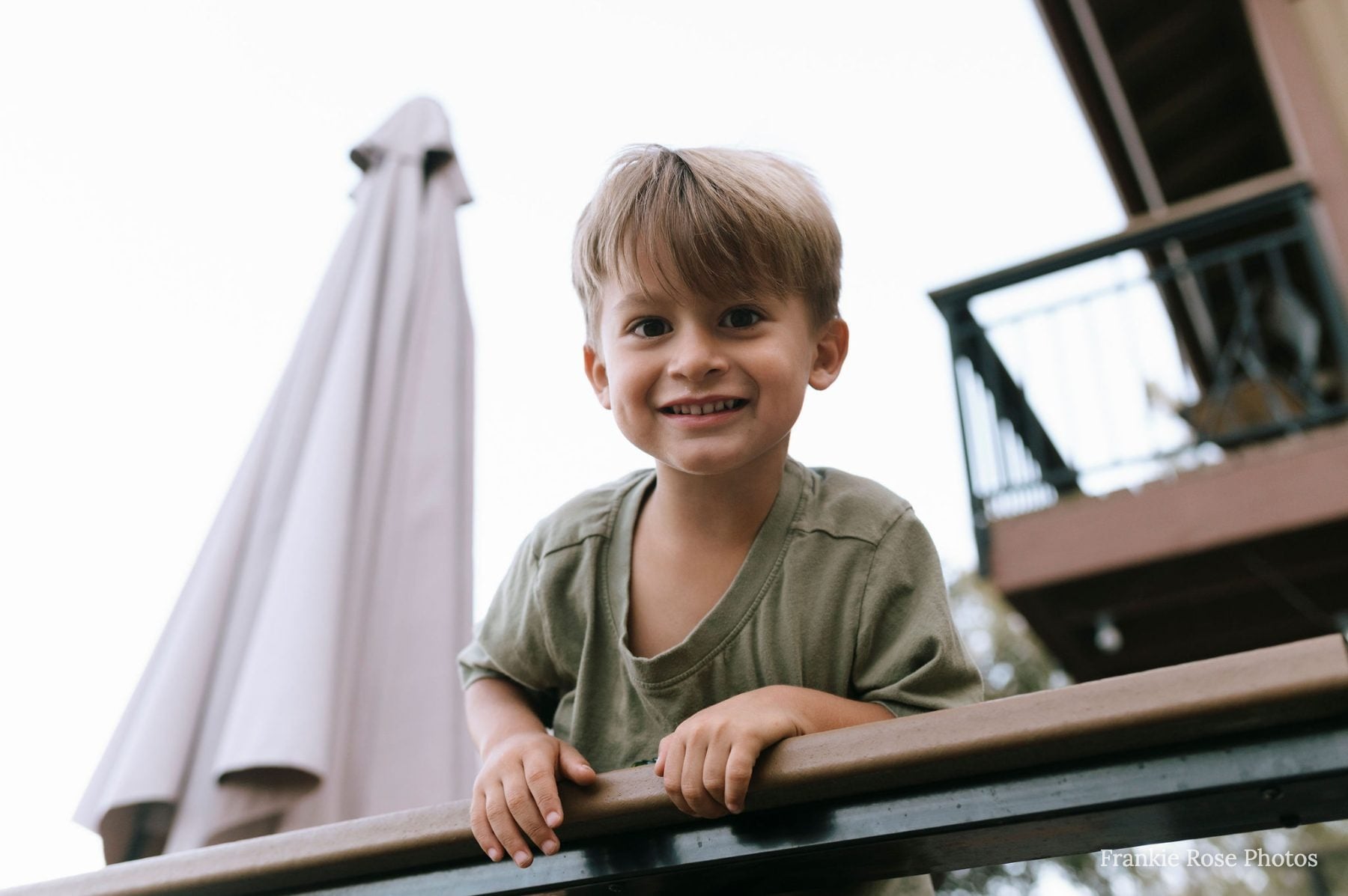 Smiling boy leans on a wooden railing near an umbrella and balcony