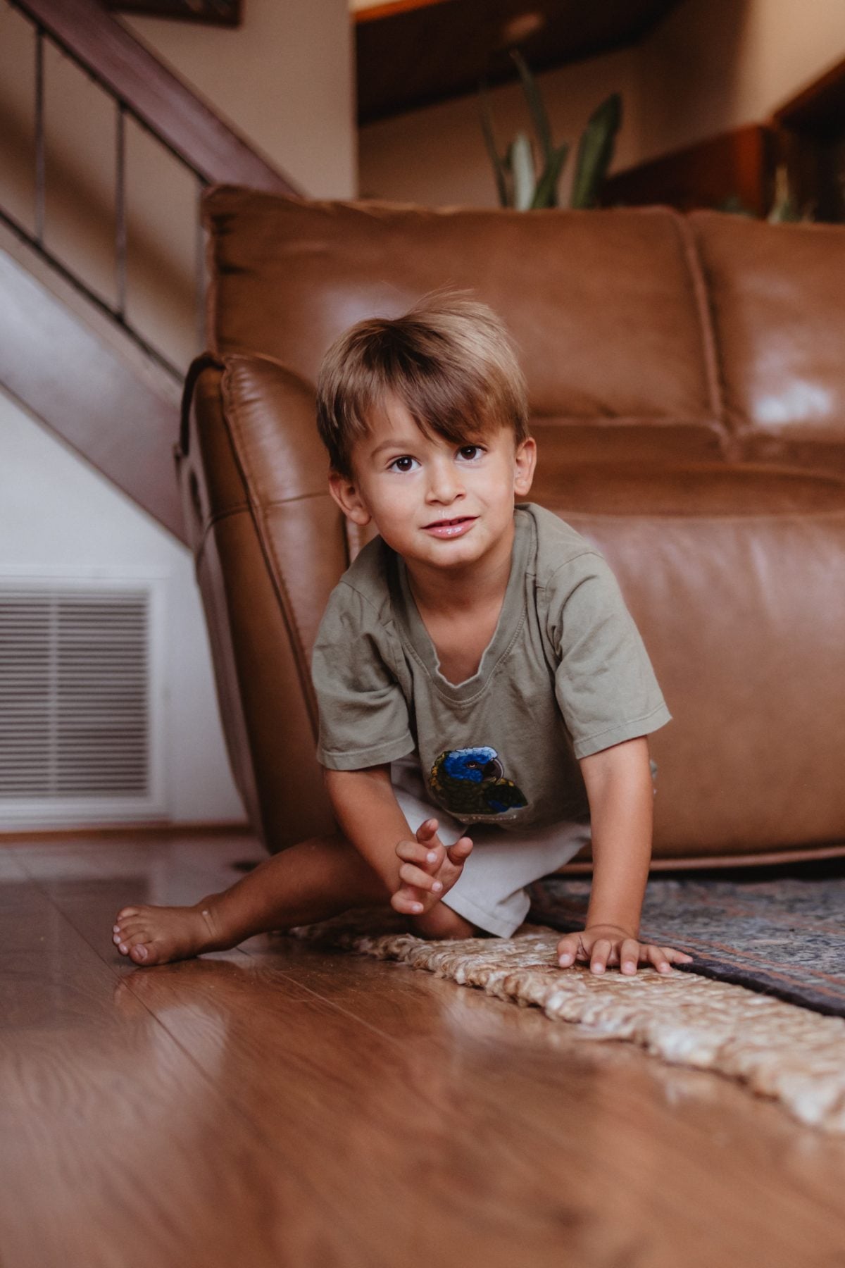 Toddler boy sits on hardwood floor beside leather couch, San Diego family photographer session