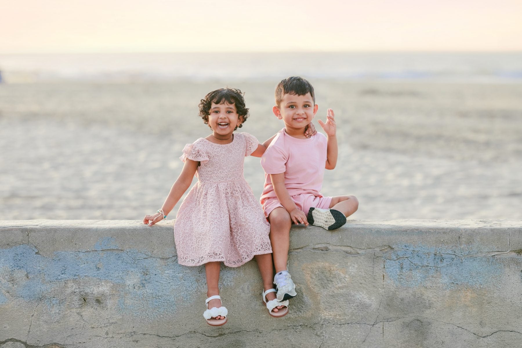 Twins in pink sit together on seawall, all smiles
