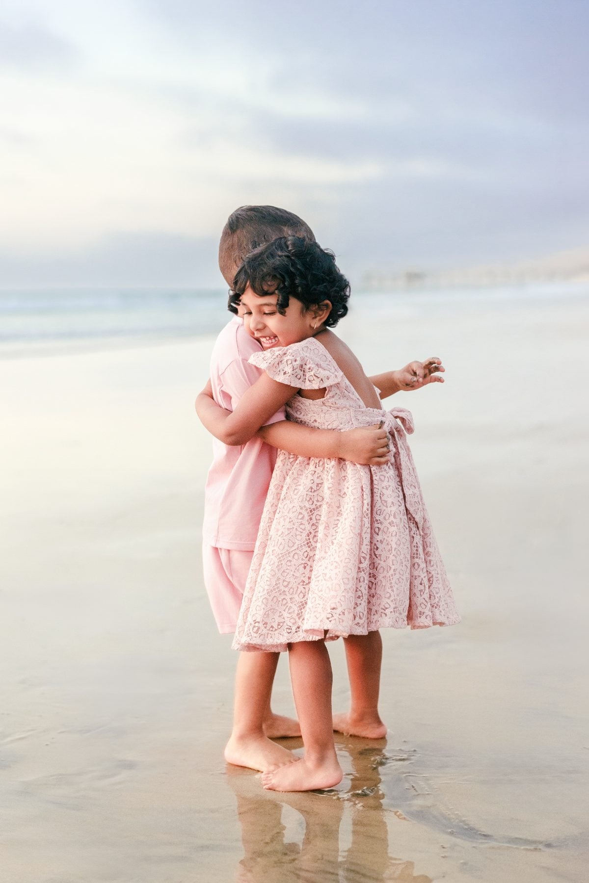 Twin toddlers in pink dresses hug each other barefoot on the sand
