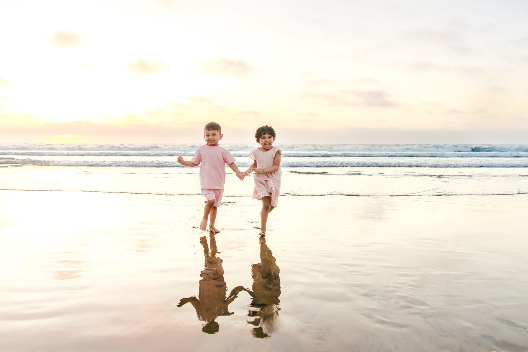 Twin siblings hold hands walking along wet sand at sunset